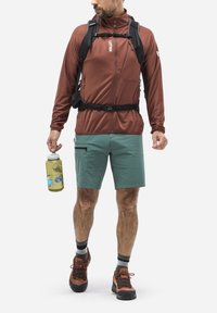 Brown zip-up jacket, green shorts, and hiking shoes. Holding a clear water bottle. Features a chest pocket and adjustable straps on the jacket.