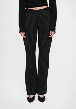 Frau trägt schwarze Nadelstreifen-Hose mit weitem Bein, schwarzes langärmeliges Cropped-Top und schwarze spitze Schuhe, steht vor einem einfarbigen Hintergrund.