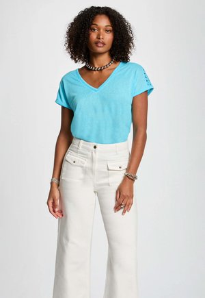 T-shirt azzurro chiaro a V con maniche corte, abbinato a pantaloni a gamba larga bianchi con tasche frontali e dettagli dorati.
