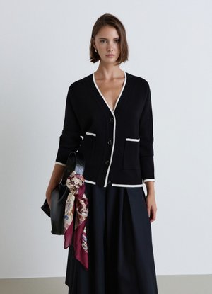 Donna che indossa un cardigan nero con bordatura bianca, gonna nera, tiene una borsa nera con una sciarpa rossa a fantasia legata alla maniglia.