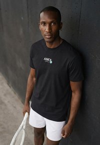 Athletischer Mann in schwarzem ASICS-Shirt und weißen Shorts lehnt an einer dunklen Wand und hält einen weißen Tennisschläger, schaut direkt nach vorne.