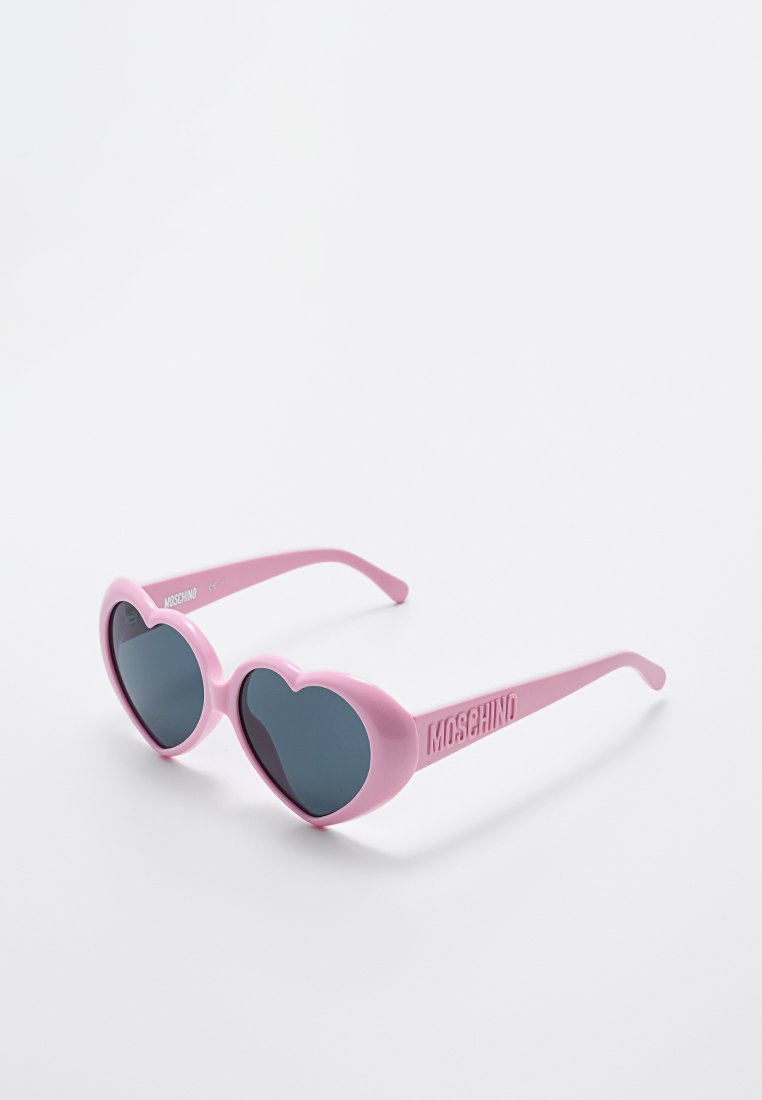 Pink heart-shaped sunglasses with dark lenses, made of plastic. Features include the brand name "Moschino" on the side of the frame.