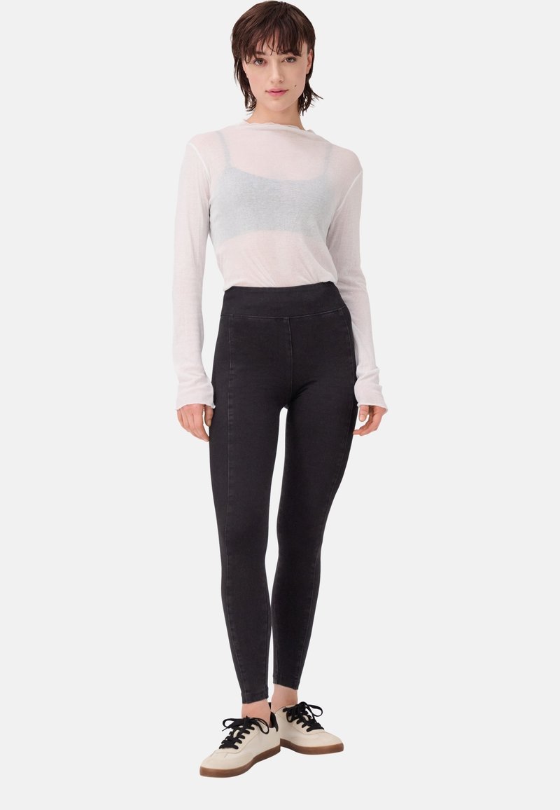 Leggings neri a vita alta realizzati in tessuto elasticizzato; abbinati a una maglietta bianca trasparente a maniche lunghe e sneaker bianche e nere.