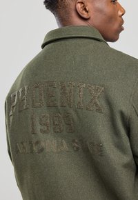 Olive-green woolen jacket featuring textured embroidery reading "PHOENIX 1969 ARIZONA STATE" on the back. Classic collar design.