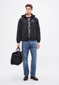 Jachetă neagră din nylon cu glugă, cu fermoar frontal și accente albastre. Asortată cu blugi albaștri și o geantă duffel neagră.