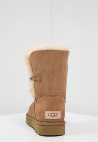 Bota de ante marrón con un suave forro de piel de crema, presenta un cierre de botón y una suela de goma plana, con el logotipo de UGG en el talón.