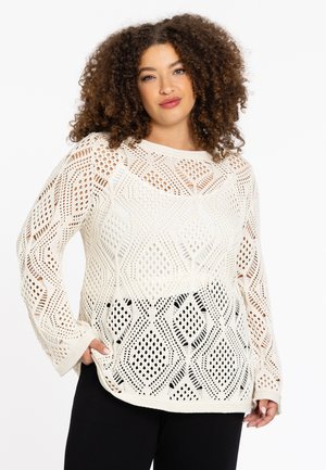 Hellbeige gestrickter Pullover mit offenen Maschenmustern, Rautenformen, langen Ärmeln und einer lockeren Passform, kombiniert mit schwarzen Hosen.