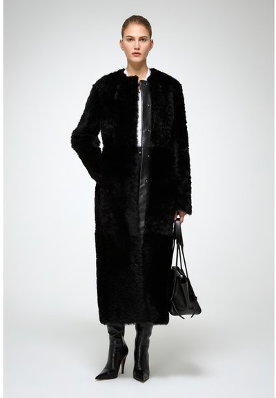 Lungo cappotto di pelliccia nera con una texture morbida, dotato di chiusura frontale e colletto rotondo. Indossato sopra un completo in pelle nera e stivali con tacco alto.