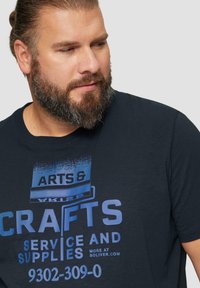 Navyblauwe T-shirt met een grafische print in verschillende tinten blauw die aan de voorkant "ARTS & CRAFTS SERVICE AND SUPPLIES" leest. Korte mouwen.