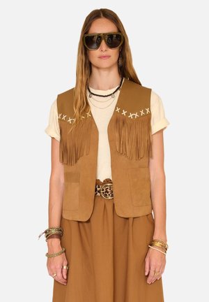 Femme portant de grandes lunettes de soleil foncées, un gilet en daim fauve à franges sur une chemise blanche aux manches retroussées, une jupe marron avec ceinture à imprimé léopard et des bracelets.