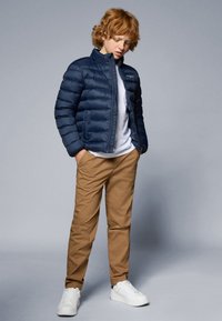 Casaco puffer azul-marinho com costuras horizontais, combinado com calças chinos bege e sapatilhas brancas. O casaco possui um colarinho alto e fecho de zip.