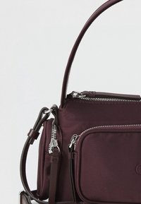 Tmavě mahagonová kabelka s hladkým tkaným povrchem; má několik kapes na zip, pevný tvar a krátkou zakřivenou rukojeť.