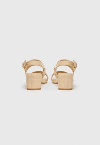 Beige high-heeled sandals with a textured fabric upper, ankle strap, and block heel, showing a woven pattern on the straps.