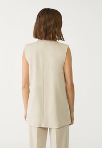 Túnica sin mangas de color beige con corte recto, textura suave y costura en la parte posterior central, diseñada para un ajuste relajado. Diseño minimalista, sin adornos.