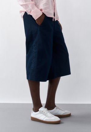 Osoba oblečená v tmavomodrých dlhých šortkách Calvin Klein, svetloružovej košeli a bielych teniskách s gumovou podrážkou, stojaca s rukami v vreckách.