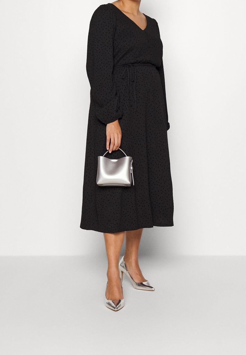 Svart omlottklänning med långa puffärmar, prickigt mönster och insvängd midja. Håller en liten silverhandväska, kompletterad med silverfärgade klackskor.