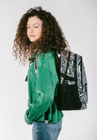 Zaino a righe bianco e nero con spallacci ergonomici, appoggiato su una giacca in suede verde. Presenta un mix di superfici lisce e testurizzate.