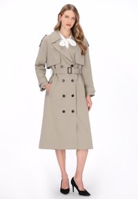 Beige trenchcoat met dubbele knoopsluiting, ceintuur in de taille, grote kraag en epauletten; gecombineerd met een witte blouse en zwarte hakken.