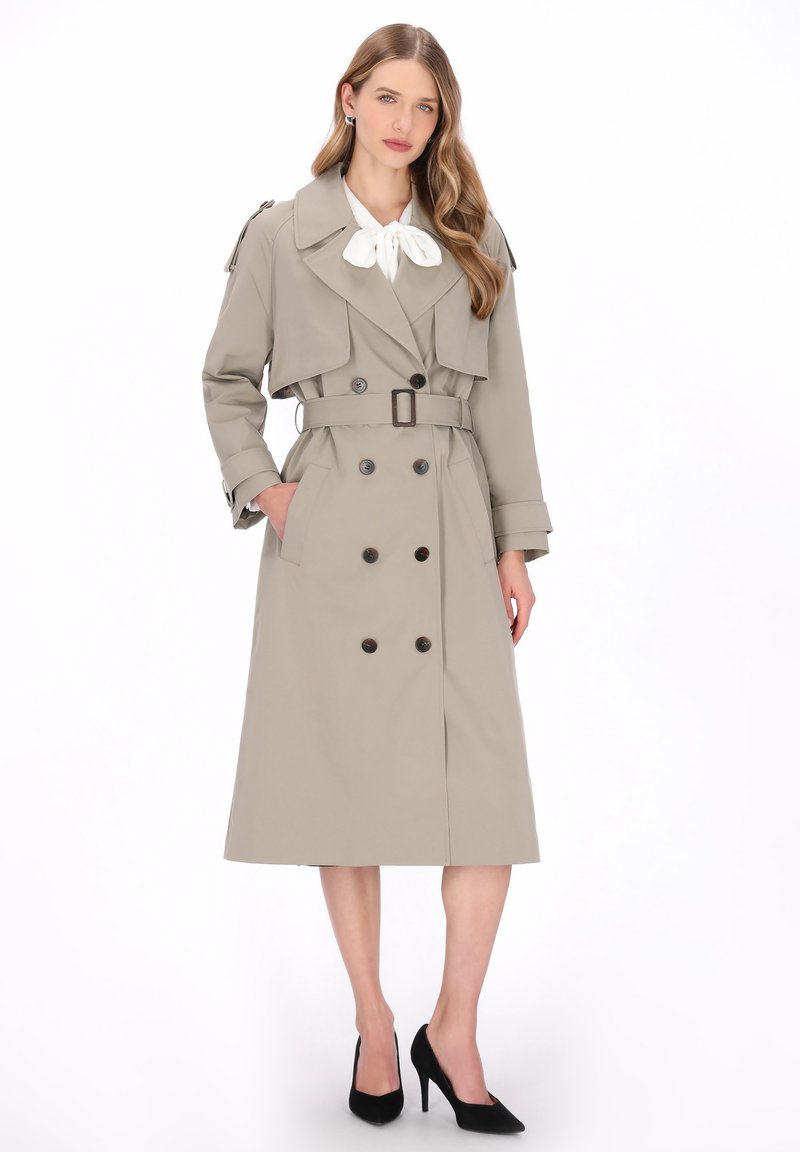 Beige trenchcoat met dubbele knoopsluiting, ceintuur in de taille, grote kraag en epauletten; gecombineerd met een witte blouse en zwarte hakken.