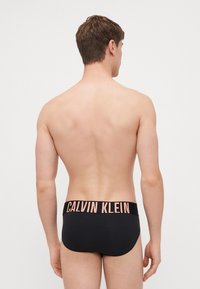 Culotte en coton noir avec une large ceinture élastique portant le logo "CALVIN KLEIN" en rose clair. Texture lisse et design ajusté.