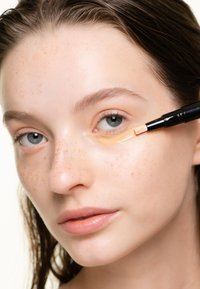 Applicatore per correttore con punta a pennello applicato su una carnagione chiara, caratterizzato da lentiggini, pelle liscia e toni neutri.