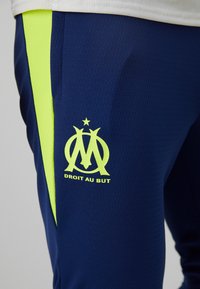 Ναυτικά αθλητικά παντελόνια με υφασμάτινη υφή, διακοσμημένα με μια φωτεινή κίτρινη λωρίδα στο πλάι και με λογότυπο που περιλαμβάνει ένα αστέρι και αναγραφή στο μηρό.