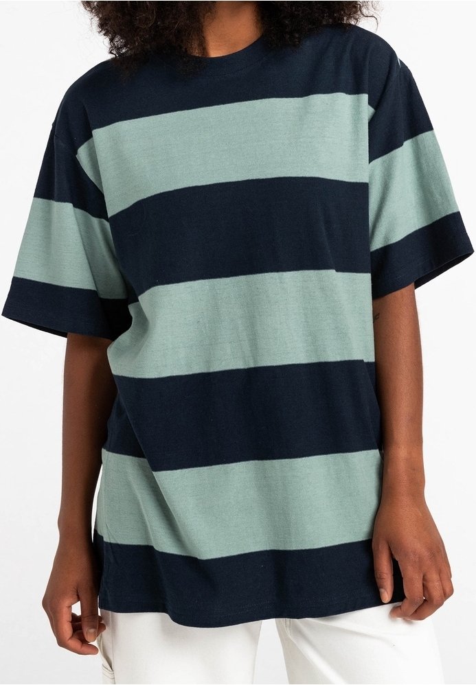 Personne portant une chemise ample à manches courtes avec de larges rayures horizontales bleu marine et vert clair, associée à un pantalon blanc.