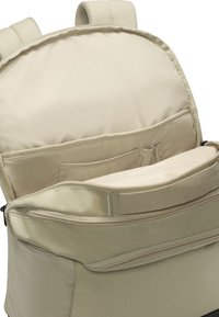 Mochila beige con un bolsillo frontal con cremallera y un sleeve de almacenamiento interior; tejido texturizado, correas acolchadas y un compartimento forrado de suave.