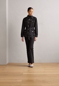 Giubbotto in denim nero con vita regolabile, chiusura a bottoni, tasche sul petto, abbinato a pantaloni neri coordinati e scarpe argentate su un pavimento di legno.
