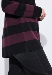 Person iført en langærmet sweater med brede vandrette sorte og mørkelilla striber samt sorte bukser med små perforeringer.