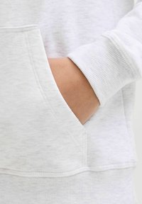 Sudadera de algodón gris con puños de canalé y un bolsillo tipo canguro. Textura suave con un ligero patrón jaspeado. Vista de cerca de una mano en el bolsillo.