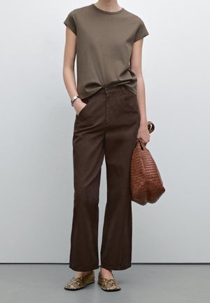 Osoba ubrana w oliwkowozielony top z krótkim rękawem, ciemnobrązowe spodnie z wysokim stanem, płaskie buty w wzór skóry węża, trzymająca dużą plecioną brązową torebkę.