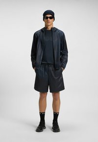 Męski strój sportowy w ciemnoniebieskim kolorze: lekkia kurtka z kapturem, dopasowane szorty, okulary przeciwsłoneczne i czarne sneakersy, zdobione subtelnymi wzorami.