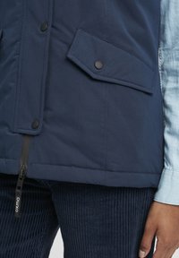 Námorně modrá vesta s zipovým uzávěrem a klopami kapsy na druky. Materiál je hladký s matným povrchem. Vnitřní vrstva je světle modrý denim.