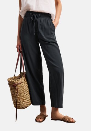 Person iført sorte bukser med snørre, hvid top, brune vævede sandaler og holder en stor flettet kurvtaske med læderstropper.