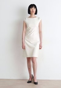 Knee-length, sleeveless cream dress with cut-out detail at shoulders, smooth texture, paired with black high heels. Simple, elegant design.