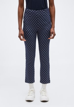 Osoba ubrana w granatowe, proste spodnie w wzory, białe skarpetki, białe trampki i czarną bluzkę, stojąca na tle jednolitego tła.