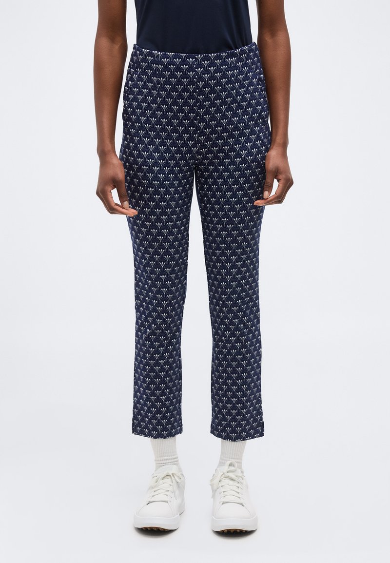 Άτομο που φοράει σκούρο μπλε παντελόνια με ευθείες μπατζάκια και σχέδιο, λευκές κάλτσες, άσπρα αθλητικά παπούτσια και μαύρο τοπ, στέκεται μπροστά σε ένα απλό φόντο.