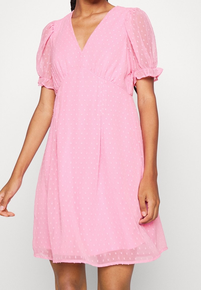 Robe rose à manches courtes avec col en V, manches bouffantes et motif à pois texturé, portée par une personne à la peau de ton moyen.