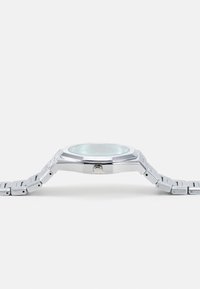 Montre en acier inoxydable argenté avec un boîtier octogonal, un cadran bleu clair et un bracelet en métal argenté. Présente un design minimaliste et une texture subtile.