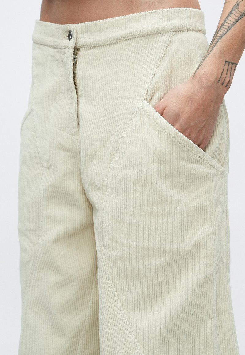 Beige Cordhosen aus Kordstoff mit Reißverschluss, Metallknopfverschluss und abgeschrägten Taschen mit gerippter Struktur.