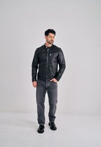 Černá kožená bunda se zipem vpředu, stojatým límcem a náprsní kapsou, doplněná šedými džíny a černými botami, na neutrálním pozadí.