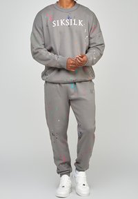 Sudadera gris y joggers a juego con manchas de pintura coloridas, con cuello redondo y puños elásticos. Texto de logo blanco en la parte delantera.