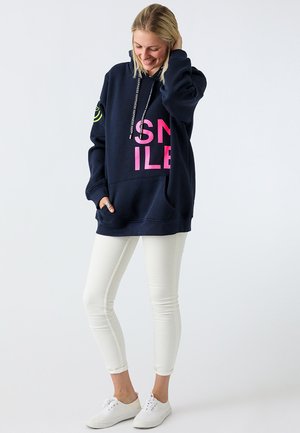 Hoodie bleu marine avec une grande impression rose "SMILE", poche frontale et capuche à cordons, assorti à un jeans skinny blanc et des baskets blanches.
