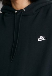 Černá mikina s kapucí vyrobená z bavlněné směsi. Má přední kapsu ve tvaru klokánka, nastavitelnou kapuci se šňůrkou a bílý logo Nike na hrudi.