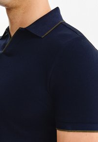 Granatowa koszulka polo z kontrastującym oliwkowozielonym kołnierzykiem i lamówką; dopasowany krój, krótkie rękawy, gładka faktura, czyste linie.