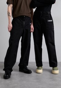 Pantalones negros con un corte relajado, que cuentan con una cremallera, bolsillos laterales y una franja reflectante. Llevados con zapatos negros voluminosos y deportivas de colores claros.