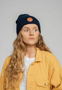 Granatowy, prążkowany czapka beanie z okrągłym pomarańczowym logo. Model nosi żółtą kurtkę na białą koszulkę, ma długie, falujące włosy.