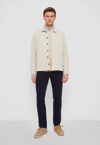 Giacca button-up in cotone beige con due tasche frontali, indossata sopra una camicia azzurra chiara, abbinata a pantaloni blu scuro e sneakers beige.