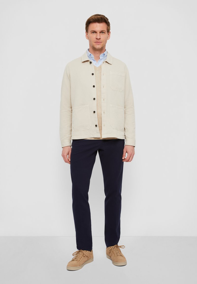 Beige Baumwollhemdjacke mit zwei Taschen auf der Vorderseite, geschichtet über einem hellblauen Hemd, kombiniert mit dunkelblauen Hosen und sandfarbenen Sneakers.
